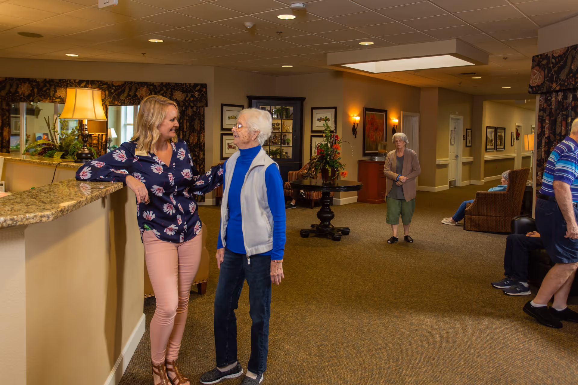 A bright and welcoming assisted living facility common area with a granite countertop reception desk on the left. A younger woman and an elderly woman are smiling and talking near the desk. In the background, other elderly residents are seated or standing in the spacious, carpeted lounge area decorated with framed artwork, plants, and warm lighting.