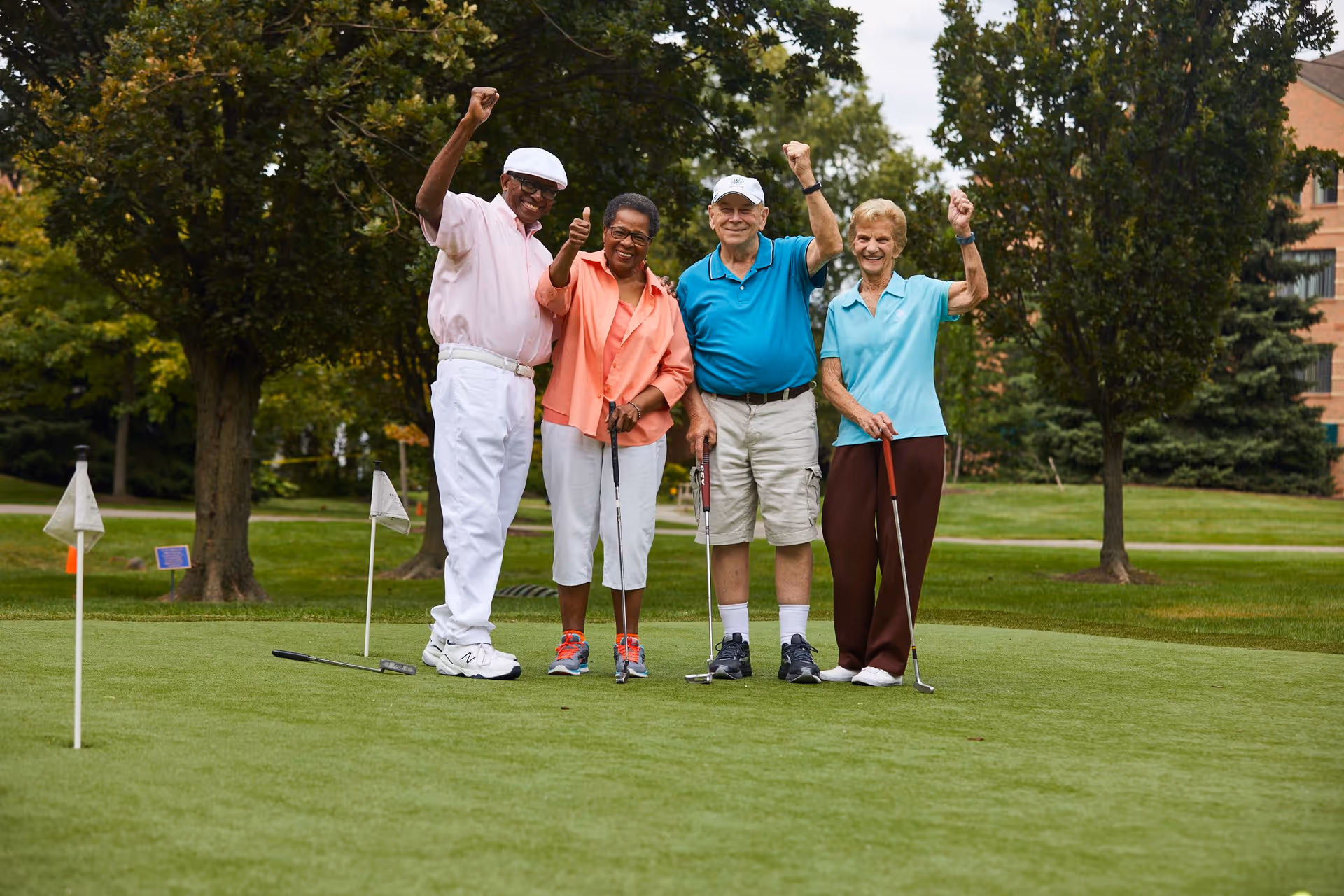 Four elderly people standing on a golf putting green outdoors, smiling and celebrating with raised arms. They are holding golf putters and dressed in casual golf attire, with trees and a building visible in the background.