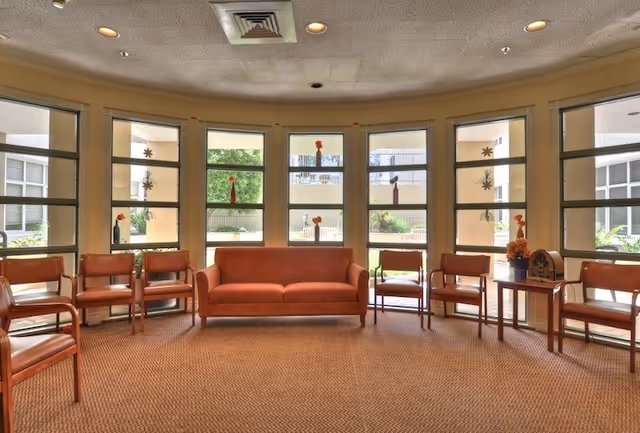 A spacious seating area with a curved arrangement of wooden chairs with brown cushions and a matching brown cushioned sofa in the center. The room has large windows with multiple panes, allowing natural light to fill the space. Small decorative items are placed on the windowsills and a side table with a flower arrangement and a decorative item is positioned to the right of the sofa. The ceiling has recessed lighting and a vent.