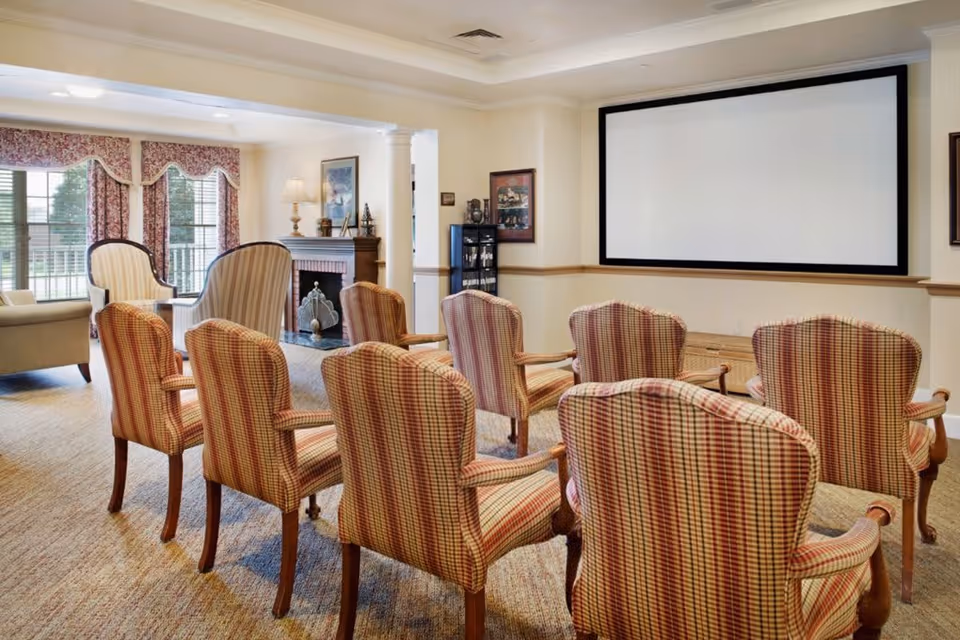 A cozy common area with several striped upholstered armchairs arranged in rows facing a large blank projection screen on the wall. The room features a fireplace with decorative items on the mantel, large windows with floral curtains, and soft lighting, creating a warm and inviting atmosphere.