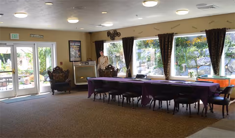 A spacious room with a long table covered with a purple tablecloth and surrounded by chairs. Large windows with dark curtains allow natural light to enter, showing a view of a rock garden outside. There are two ornate chairs near a glass door leading outside, and a person standing near the windows. The ceiling has multiple round lights.