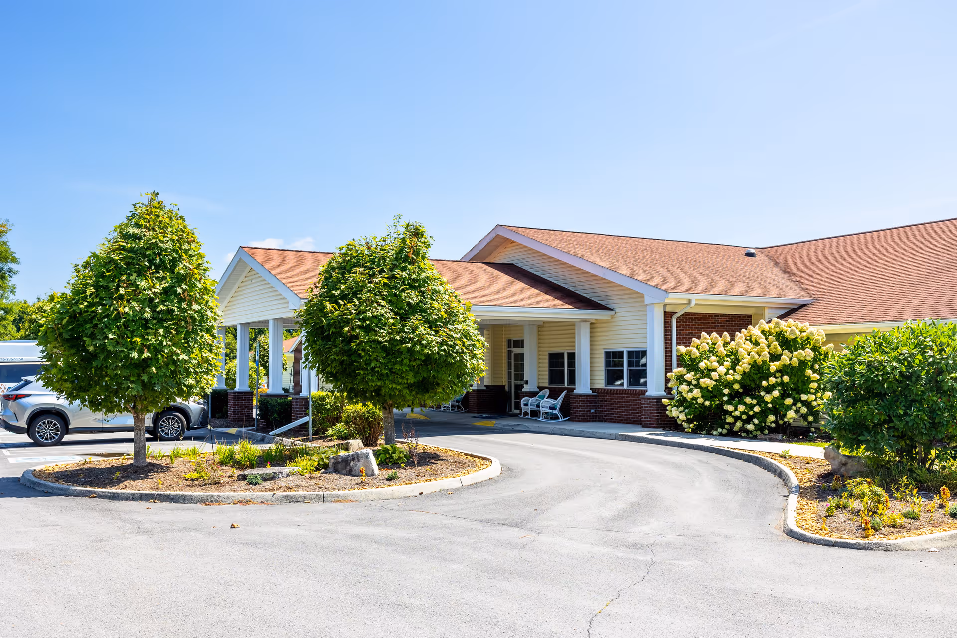 Exterior view of Commonwealth Memory Care at Cedar Bluff facility showing a single-story building with a covered entrance, surrounded by landscaped greenery including trees and flowering bushes, under a clear blue sky.