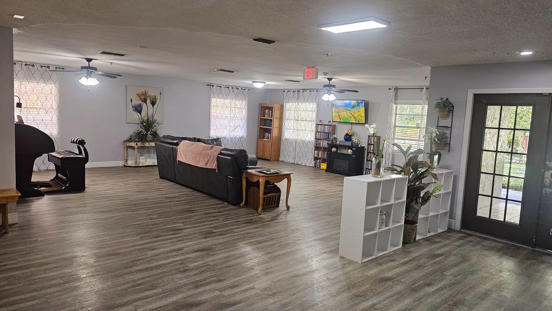 Spacious communal living room with sofas, TV, shelving, plants and a glass door in an assisted living facility.