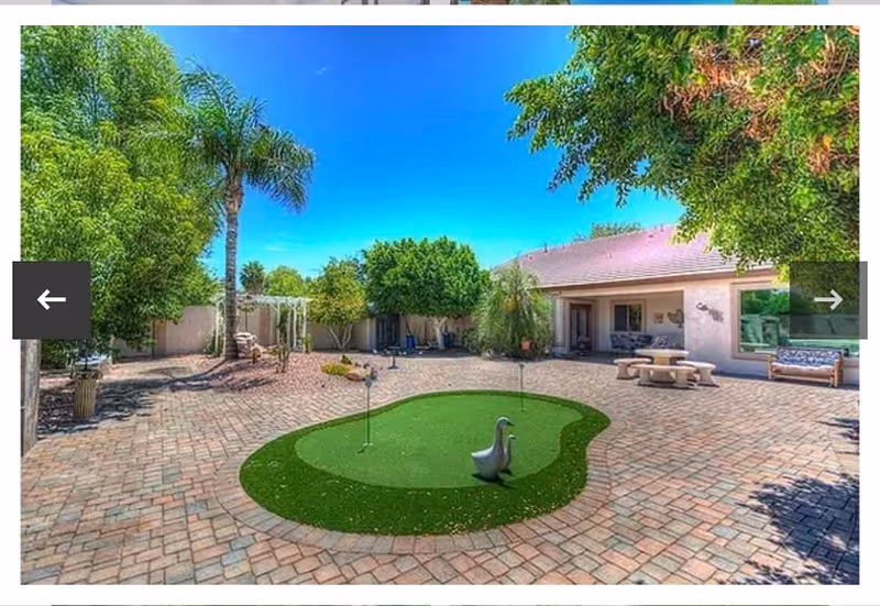 Outdoor courtyard area with a small artificial putting green shaped like a pond with a duck decoration, surrounded by brick pavers. There are trees and shrubs around the perimeter, a picnic table with benches, and a bench near the building. The sky is clear and blue.