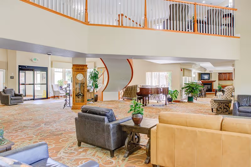 Spacious senior living facility common area with a high ceiling and upper balcony. The room features various seating options including leather and fabric armchairs and sofas arranged around wooden side tables with potted plants. A grand piano is positioned near a curved staircase in the background. Large windows allow natural light to fill the space, and there is a grandfather clock near the entrance door.