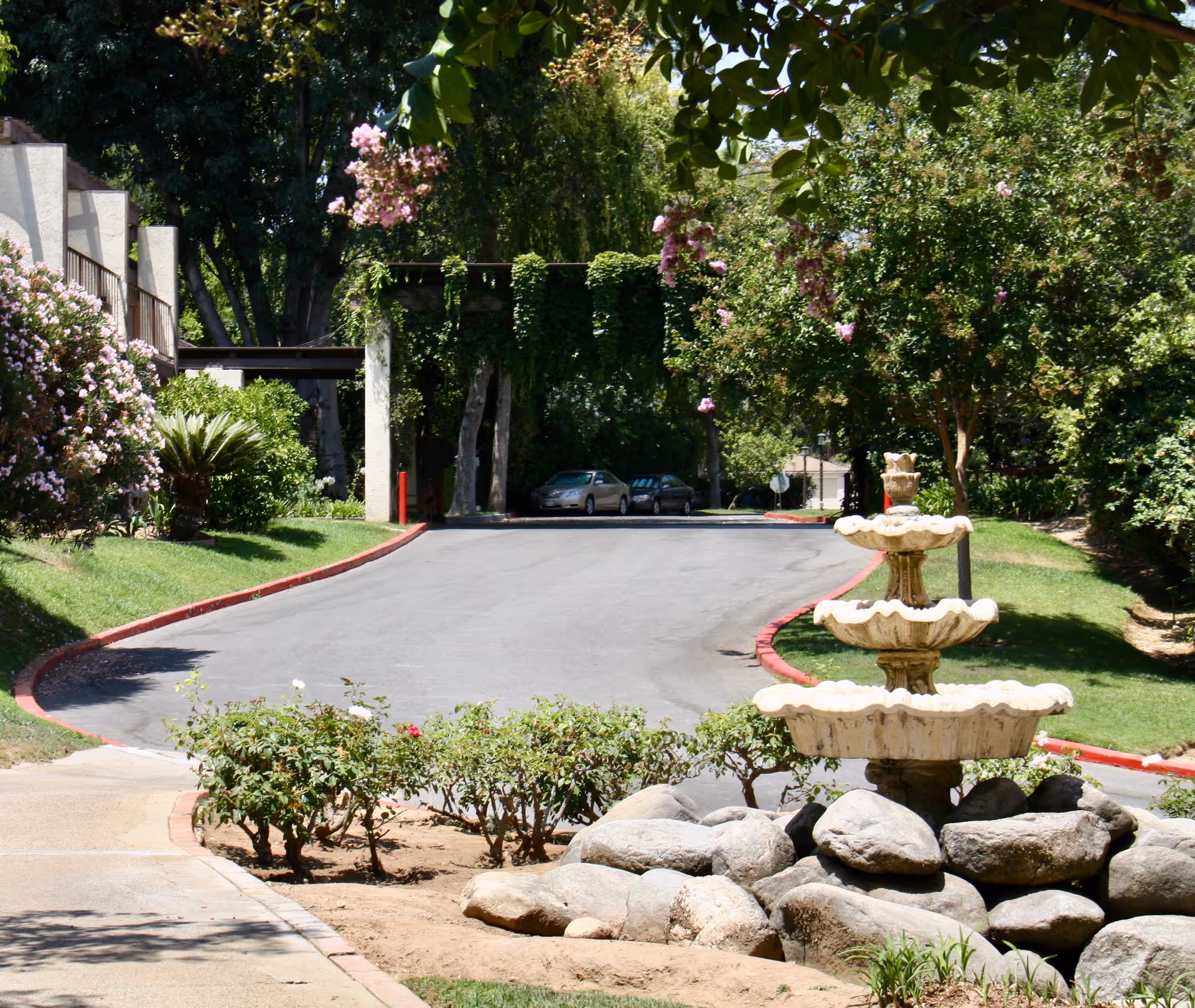 Outdoor view of a driveway lined with greenery and flowering bushes, leading to a shaded area with parked cars. In the foreground, there is a three-tiered stone fountain surrounded by rocks and plants.