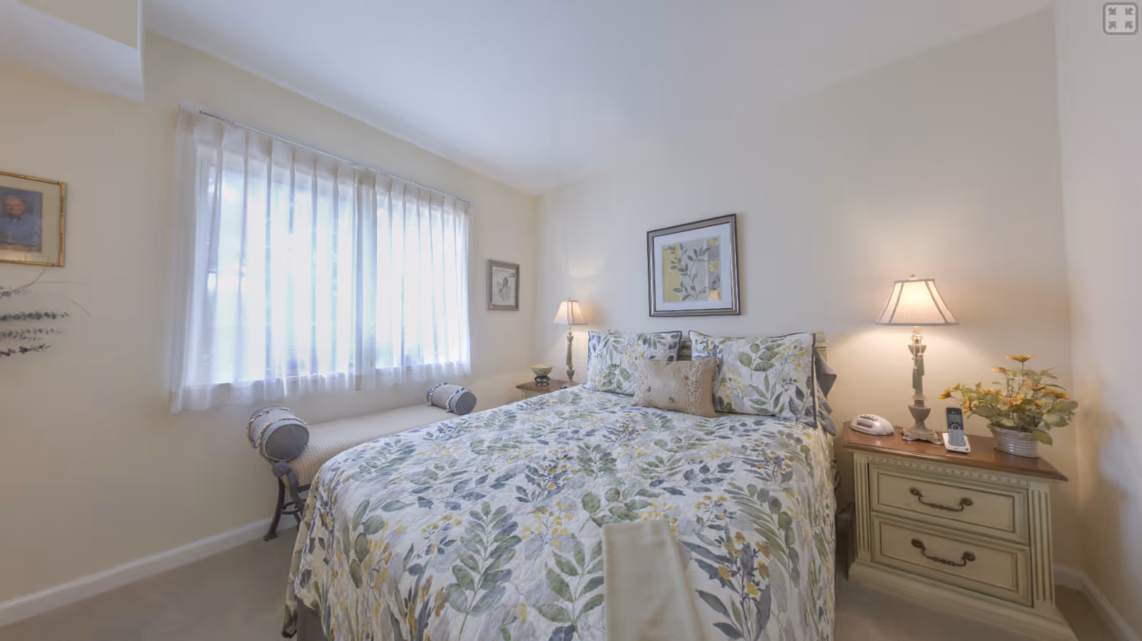 A cozy bedroom with a bed covered in a floral patterned quilt and matching pillows. There are two bedside tables with lamps, a telephone, and a small plant. A window with sheer white curtains lets in natural light, and framed artwork hangs on the walls.