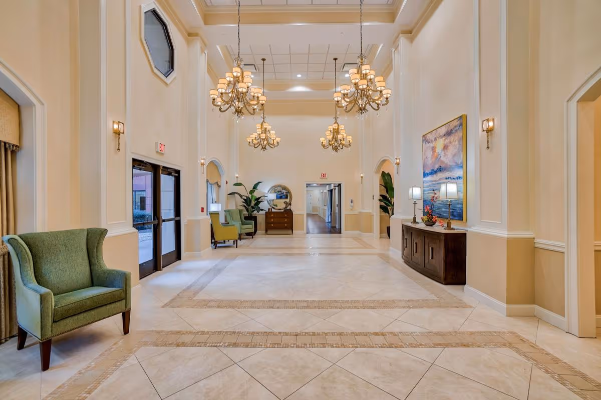 Spacious and elegant interior hallway with high ceilings, multiple chandeliers, beige tiled floor with decorative borders, green upholstered chairs along the walls, a wooden console table with lamps and a large colorful painting, and double glass doors leading outside.