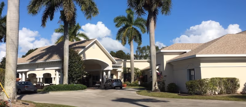 Exterior front entrance of a single-story senior living building with a covered driveway, palm trees, and parked cars.