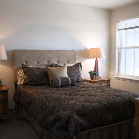 A neatly made bed with a tufted beige headboard, dark patterned bedding, and several pillows. There are wooden nightstands on either side of the bed, each with a lamp and a small plant on the right nightstand. A window with blinds is visible on the right side of the room, allowing natural light to enter.