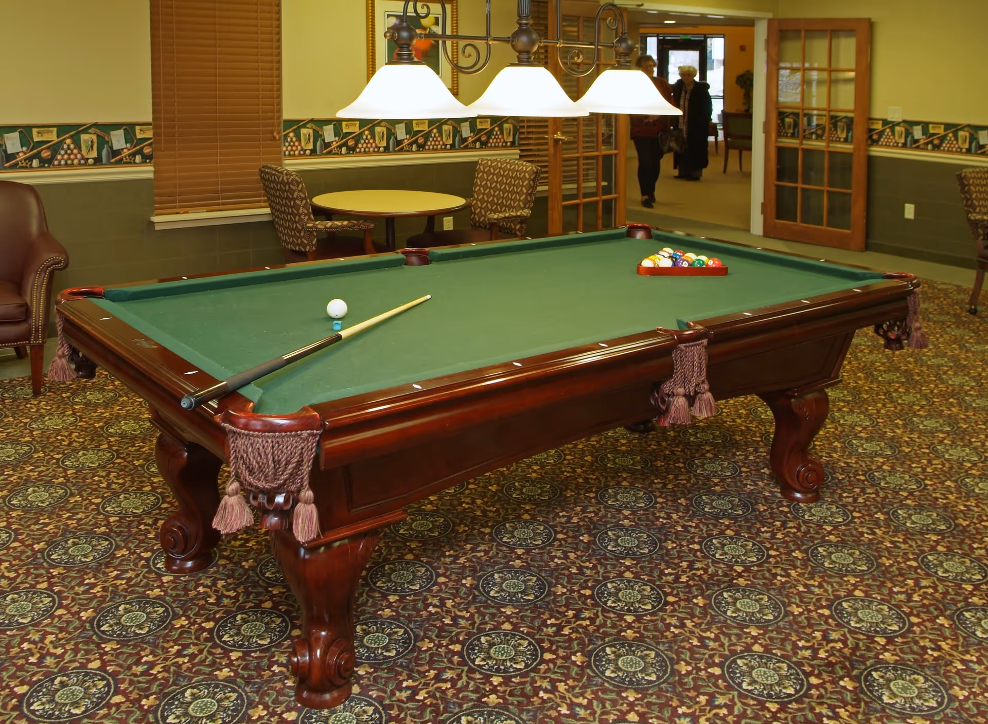 A billiards room with a green felt pool table, a cue stick resting on the table, and a rack of pool balls arranged for a game. The room has patterned carpet, a round table with two chairs, a leather armchair, and a hanging light fixture above the pool table. In the background, there are two elderly women walking through a doorway with glass-paneled wooden doors.
