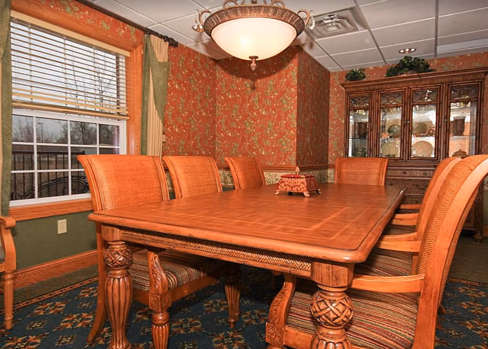 A formal dining room with a large wooden table surrounded by wicker-backed chairs, patterned wallpaper, a china cabinet, and a window.