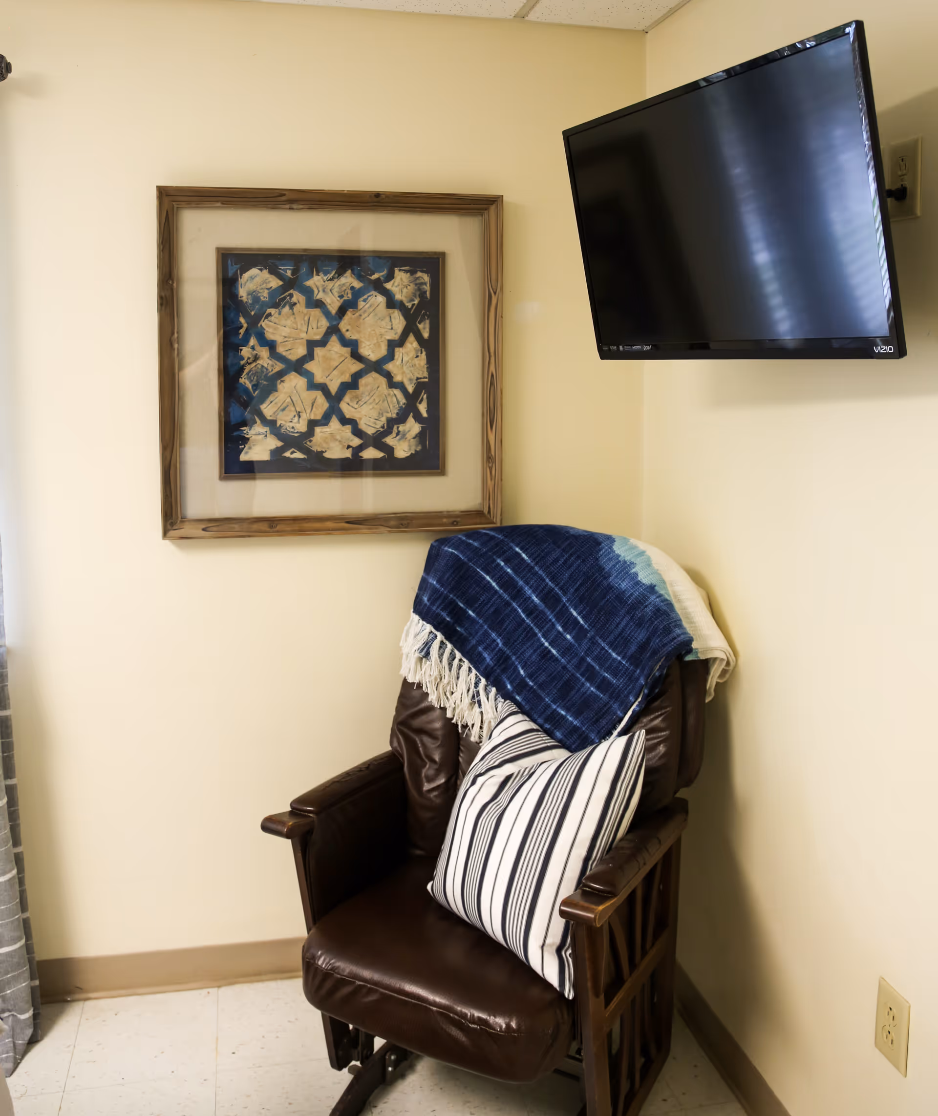 A cozy corner with a brown leather armchair featuring wooden armrests, a striped pillow, and a blue and white throw blanket draped over the back. Above the chair is a framed abstract artwork with a wooden frame. A flat-screen Vizio TV is mounted on the adjacent wall. The walls are painted light yellow and the floor is tiled.