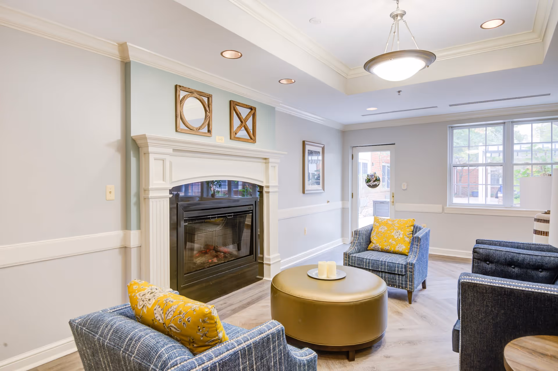 A cozy senior living facility common area with a white fireplace, two blue armchairs with yellow floral pillows, a round gold ottoman with candles on a tray, large windows letting in natural light, and a door with a welcome sign.