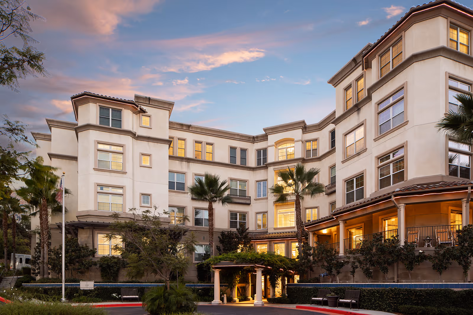 Exterior view of a multi-story senior living facility building at dusk with warm lights glowing from the windows, palm trees, and a covered entrance area with benches and landscaping.