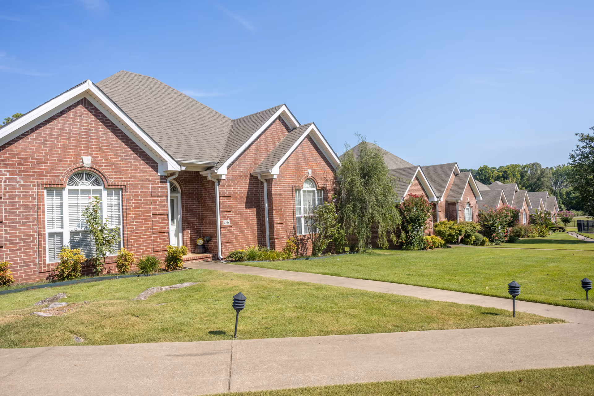 Row of single-story red brick residential units with manicured lawns and a sidewalk under a clear blue sky.