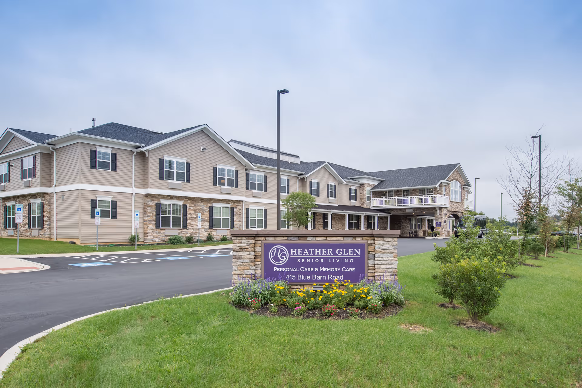 Exterior view of Heather Glen Senior Living facility showing a two-story building with beige siding and stone accents. There is a purple sign in front with the facility name and address, surrounded by a flower bed and green lawn. The parking lot includes several handicap parking spaces.