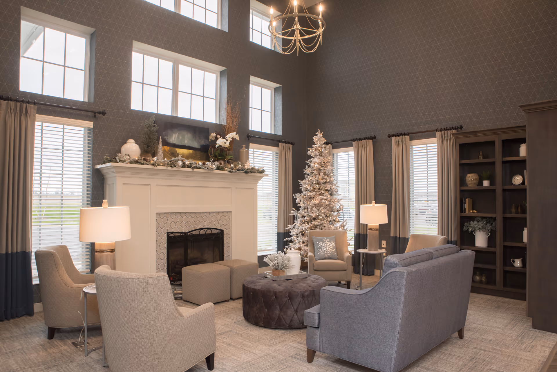A cozy senior living facility common area with high ceilings and large windows. The room features a decorated white fireplace mantel with holiday greenery and ornaments, a white Christmas tree with lights, beige and gray armchairs, a gray sofa, two side tables with lamps, a round tufted ottoman, and a dark wood bookshelf with decorative items.