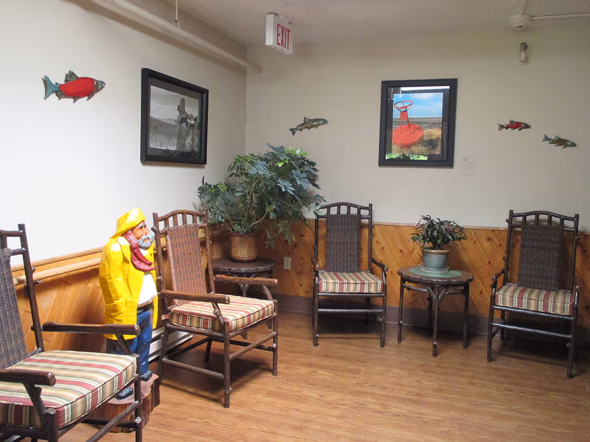 A small seating area with four wooden chairs featuring striped cushions arranged against a wall with wood paneling. There is a small round table with a potted plant on it and a larger potted plant in the corner. The walls are decorated with framed pictures and fish-shaped wall hangings. A statue of a fisherman wearing a yellow raincoat and hat stands near one of the chairs. An exit sign is visible above the seating area.
