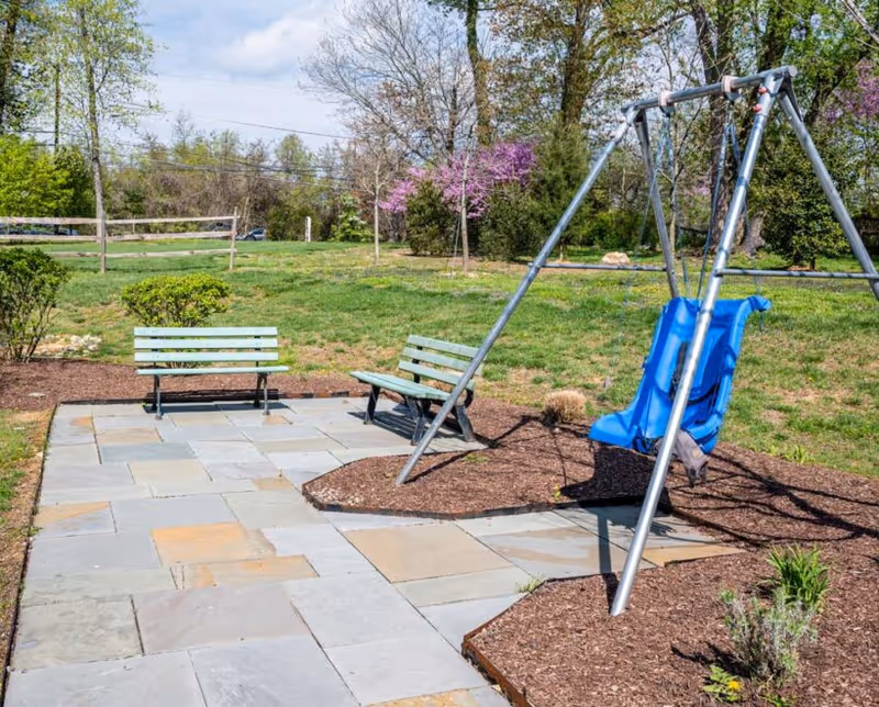 Outdoor area with a paved stone walkway, two green benches, and a blue swing set on a mulched patch surrounded by grass and trees with some blooming flowers in the background.