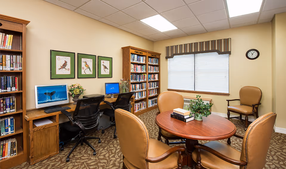 A cozy library or reading room with wooden bookshelves filled with books, two computer workstations with black mesh chairs, a round wooden table with four tan upholstered chairs, a small plant and books on the table, three framed bird illustrations on the wall, a window with blinds and a striped valance, and a wall clock.