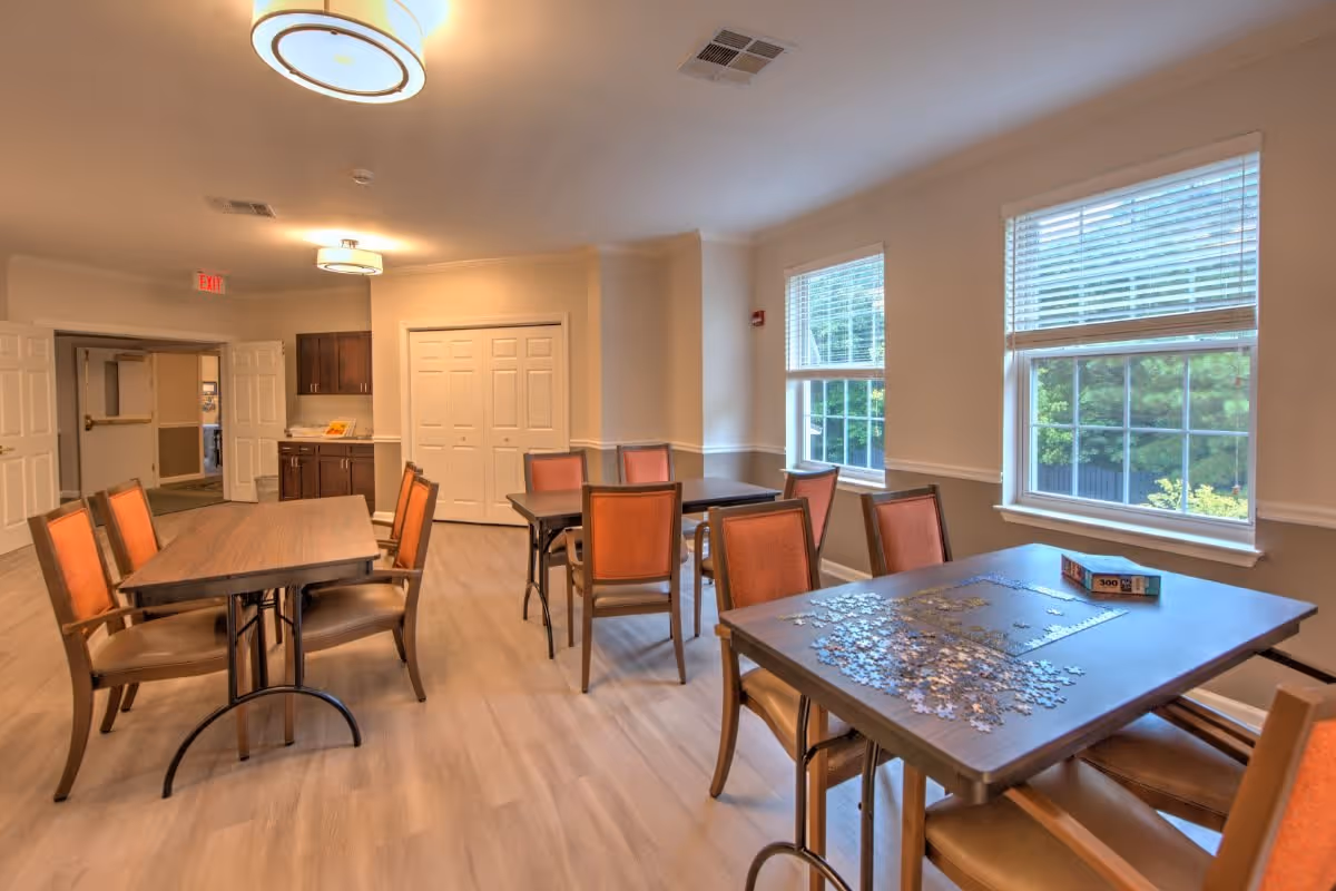 A well-lit room with multiple tables and chairs arranged for group activities. One table near the window has a partially completed jigsaw puzzle and a puzzle box on it. The room has large windows with blinds, light-colored walls, and wooden flooring. There are ceiling lights and a cabinet with a countertop in the background.