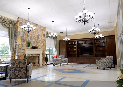 A spacious and well-lit living room area with patterned armchairs arranged around a stone fireplace. The room features large windows with curtains, multiple chandeliers hanging from the ceiling, and a large wooden entertainment center with a flat-screen TV and decorative items.