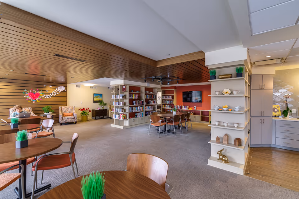A spacious and well-lit common area in a senior living facility featuring round wooden tables with chairs, small green plant decorations on the tables, a bookshelf filled with books, a wall-mounted TV, and comfortable seating including armchairs. The ceiling has wooden paneling and the room has a cozy, inviting atmosphere with decorative elements such as flowers and small sculptures.