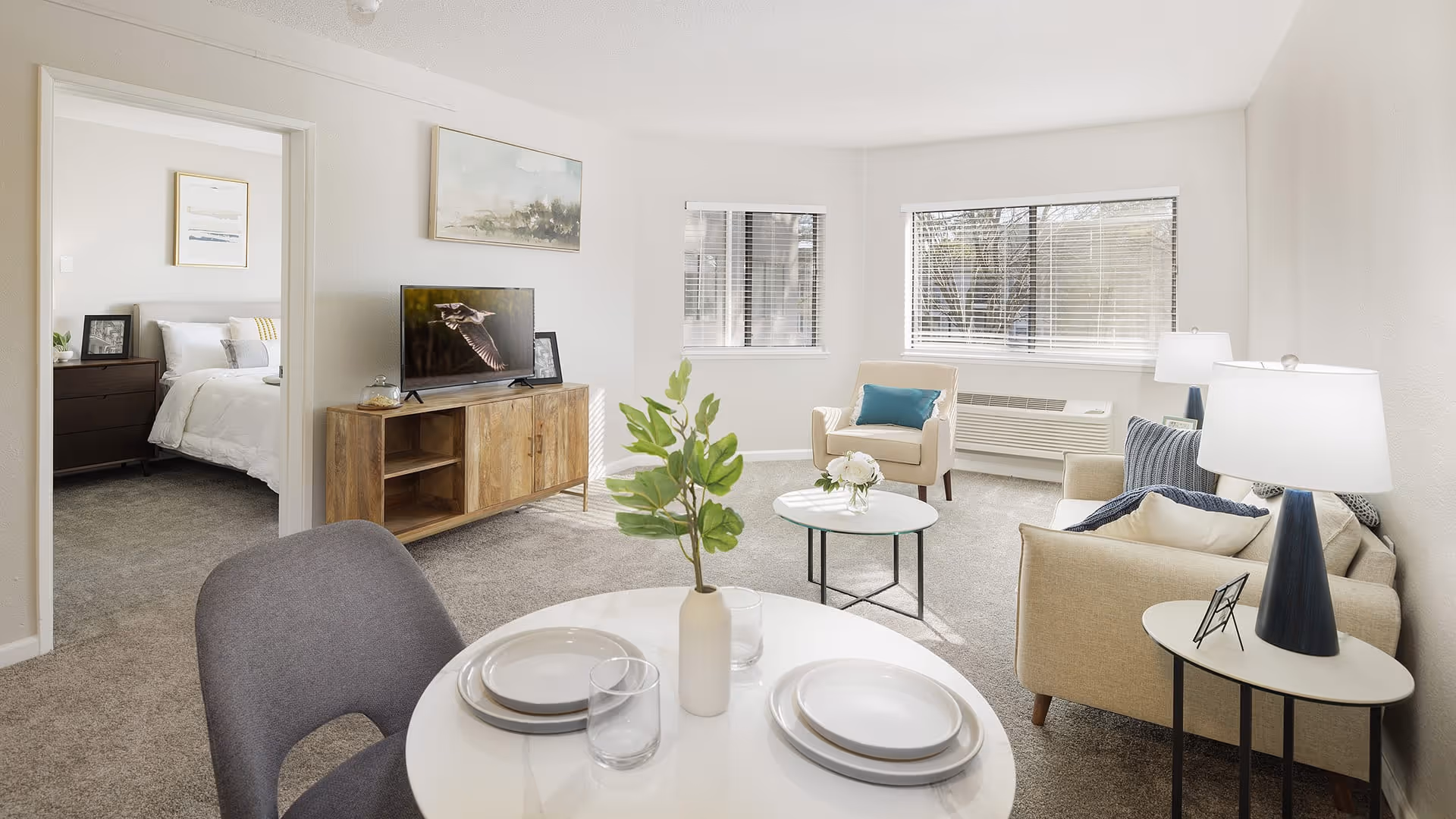 A bright and cozy living room with beige walls and carpeted floor. The room features a beige sofa with cushions, a beige armchair with a blue pillow, a round glass coffee table with a small flower arrangement, and a wooden TV stand with a flat-screen TV. A round dining table with two place settings and a vase with green leaves is in the foreground. Two windows with blinds allow natural light to fill the room. A bedroom with a neatly made bed and framed artwork is visible through an open doorway.