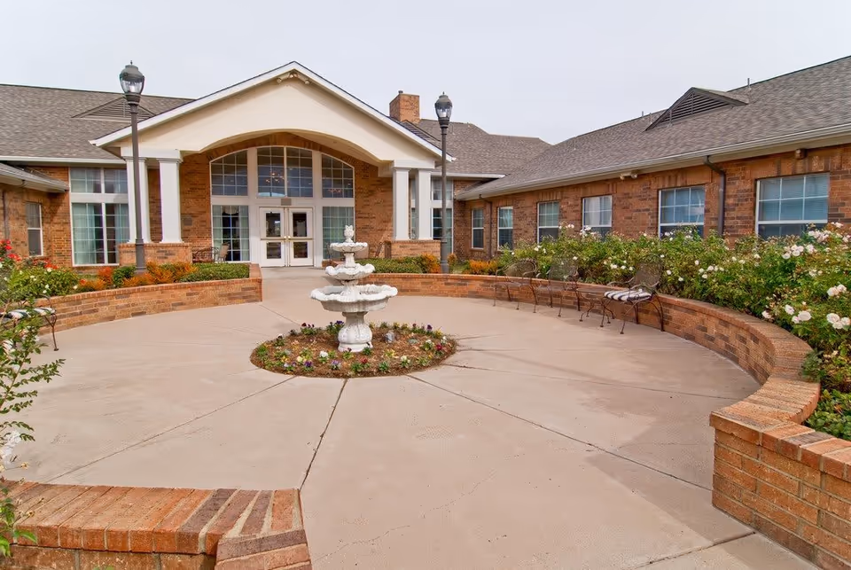 Front exterior view of Morada Cedar Hill Assisted Living and Memory Care building with a circular paved area featuring a white tiered fountain in the center, surrounded by flower beds and benches. The building has large windows and a covered entrance with white columns.
