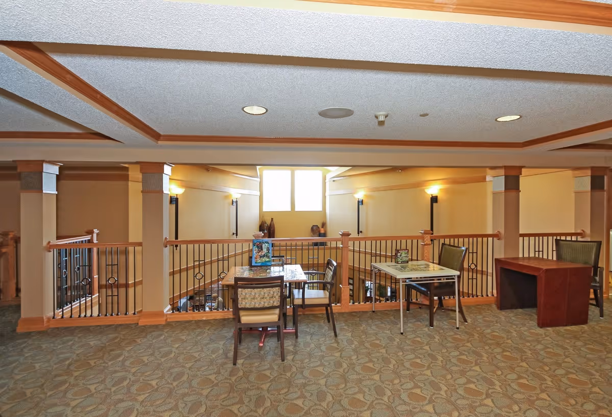 Interior view of a senior living facility area with carpeted floor, two small tables each with two chairs, a wooden desk with a chair, and a railing overlooking a lower level. The walls are painted beige with wall-mounted lights and a large window letting in natural light.