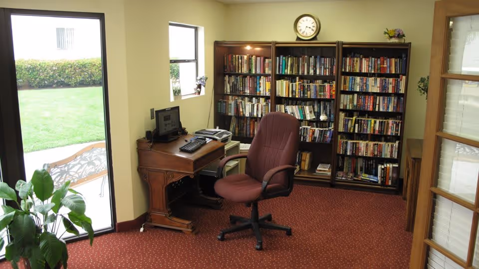 A cozy reading and computer area in a senior living facility with a wooden desk holding a computer and printer, a maroon office chair, a large bookshelf filled with books, a clock on the wall above the bookshelf, a window, a glass door leading outside to a garden with a bench, and a green plant near the door.