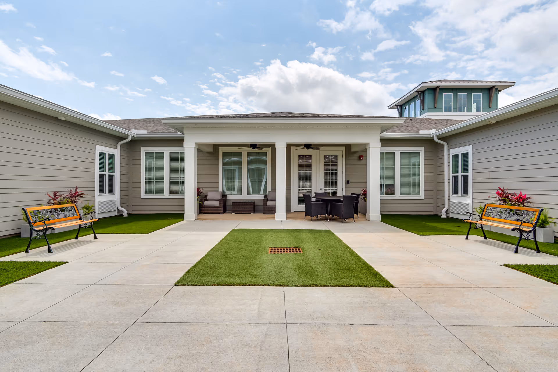 Outdoor courtyard area at The Canopy at Warner Robins featuring a concrete patio with green artificial grass patches, two wooden benches with black metal frames, potted plants with red and green foliage, and a covered seating area with cushioned chairs and a table under a roof supported by white columns.
