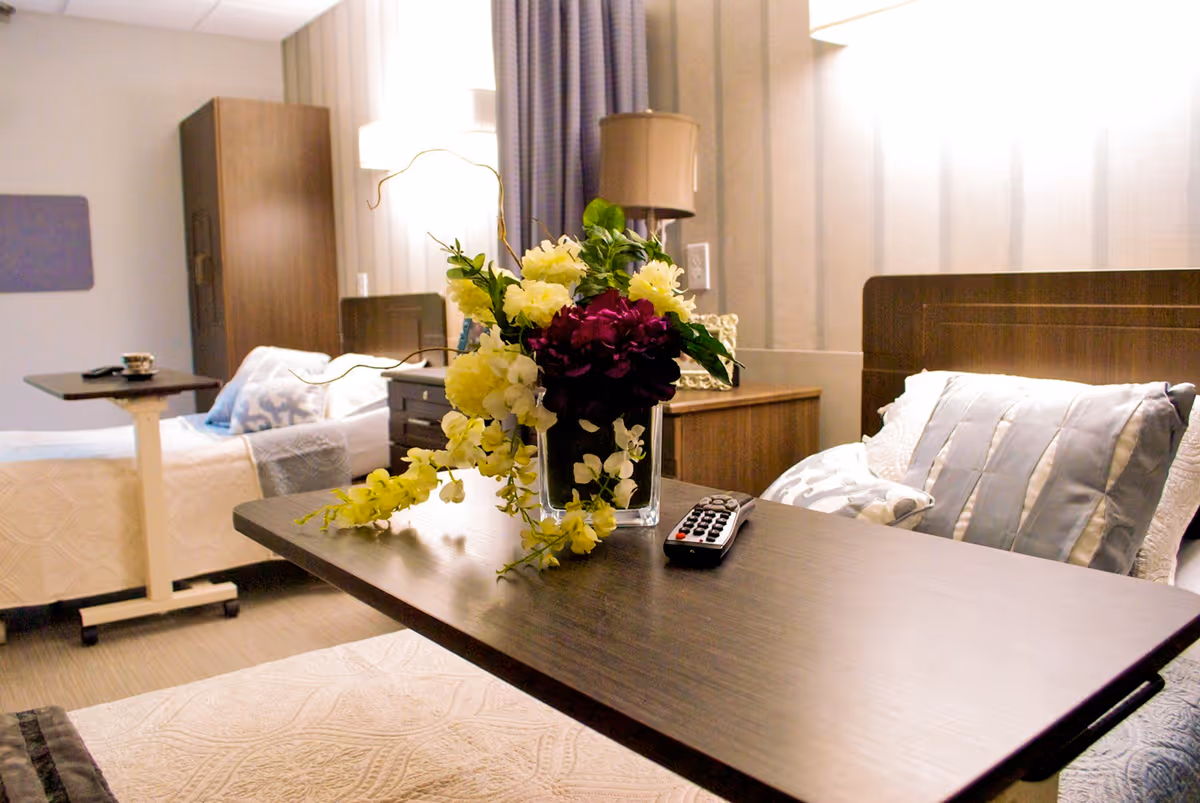 A well-lit bedroom in a senior living facility featuring two beds with pillows and blankets. A dark wooden overbed table is positioned over one bed, holding a vase with a colorful flower arrangement and a TV remote control. In the background, there is a wooden wardrobe, a nightstand with a lamp, and curtains covering the window.