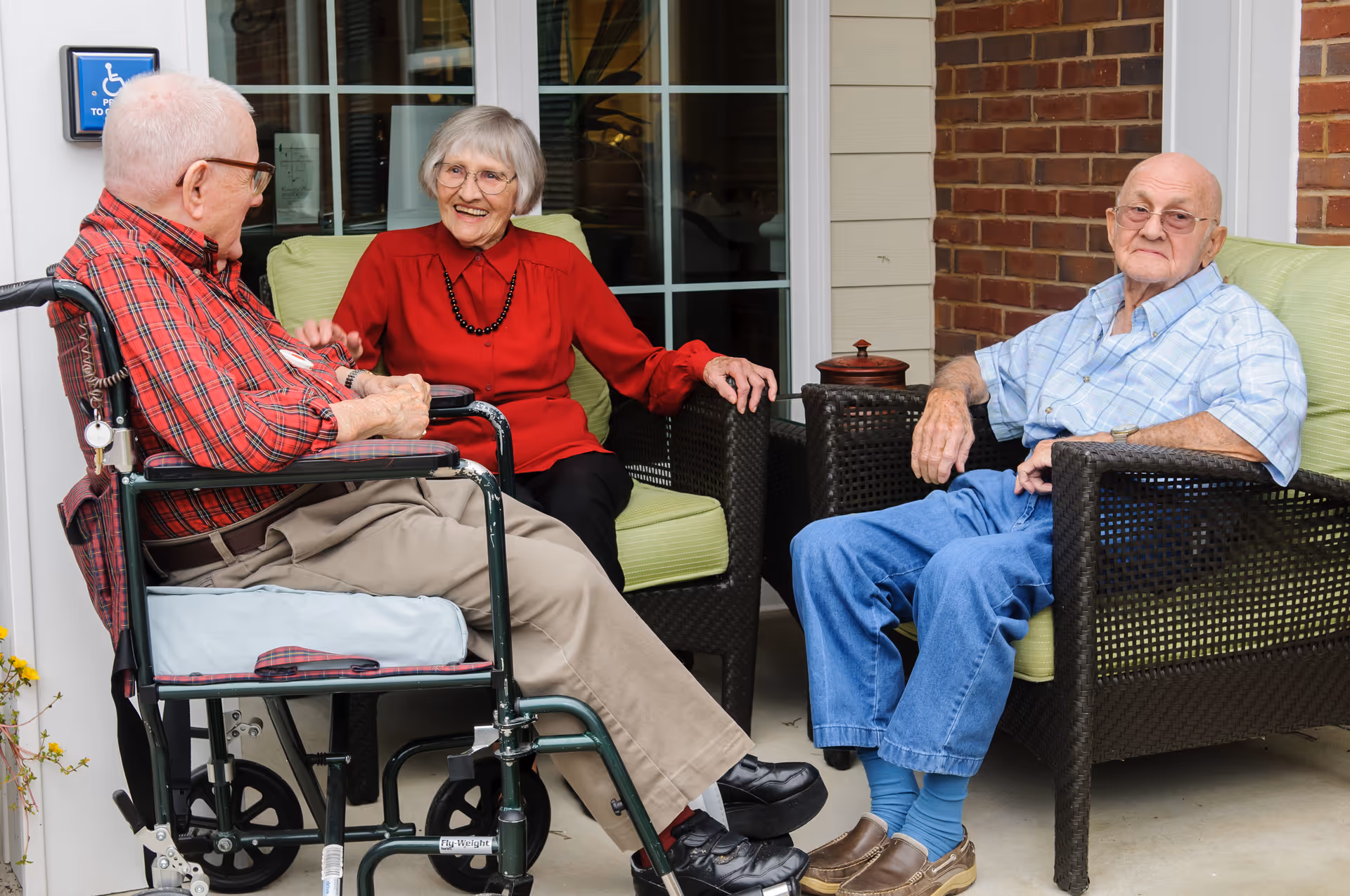 Three elderly individuals sitting and chatting on a patio outside a building. One man in a wheelchair wearing a red plaid shirt and beige pants, a woman in a red blouse and black pants sitting on a green cushioned chair, and another man in a blue plaid shirt and blue pants sitting on a similar chair. The background shows a window and brick wall.
