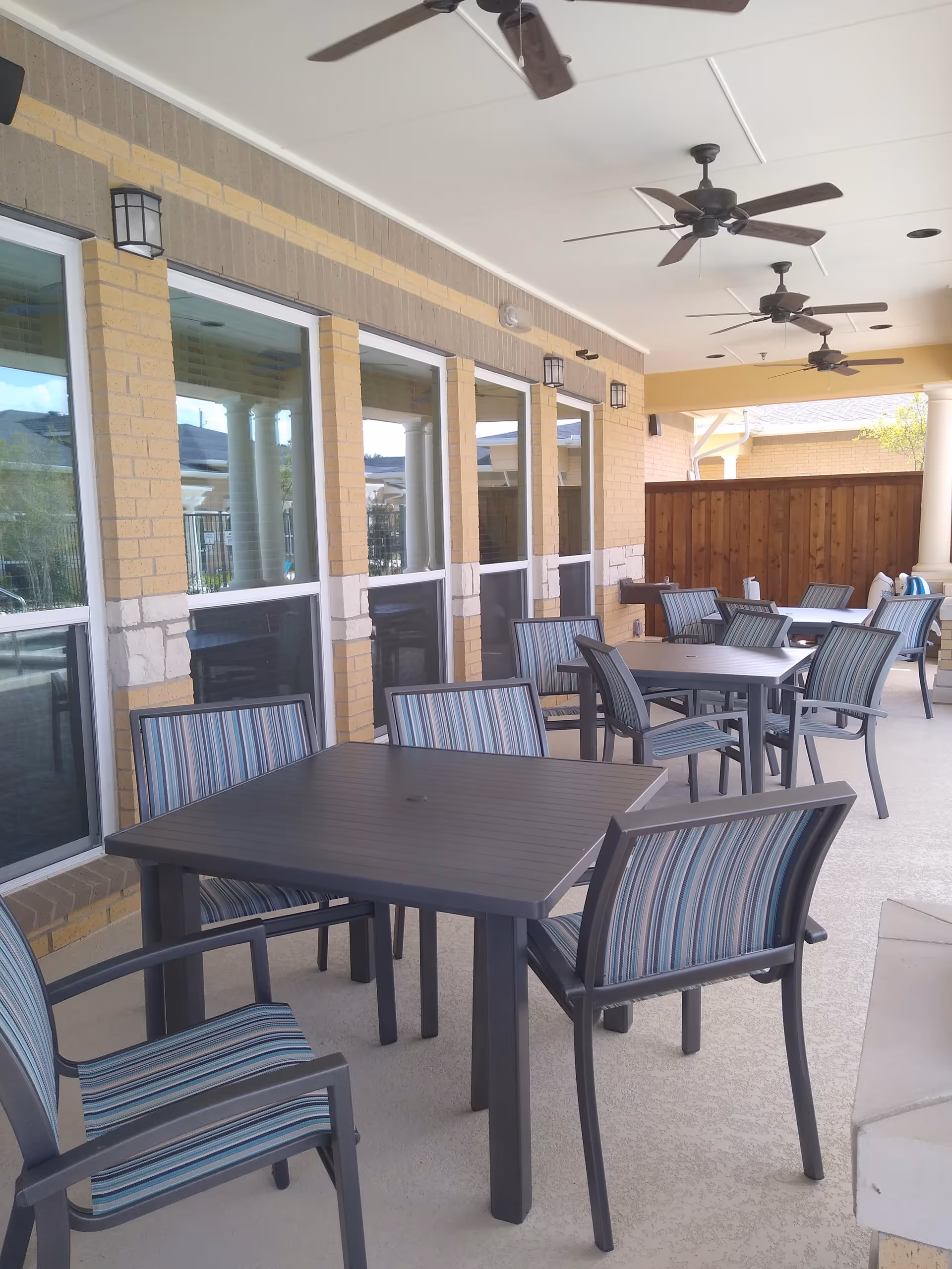 Outdoor covered patio area with multiple square tables and striped cushioned chairs. The patio has ceiling fans and is adjacent to a building with large windows and brick walls.