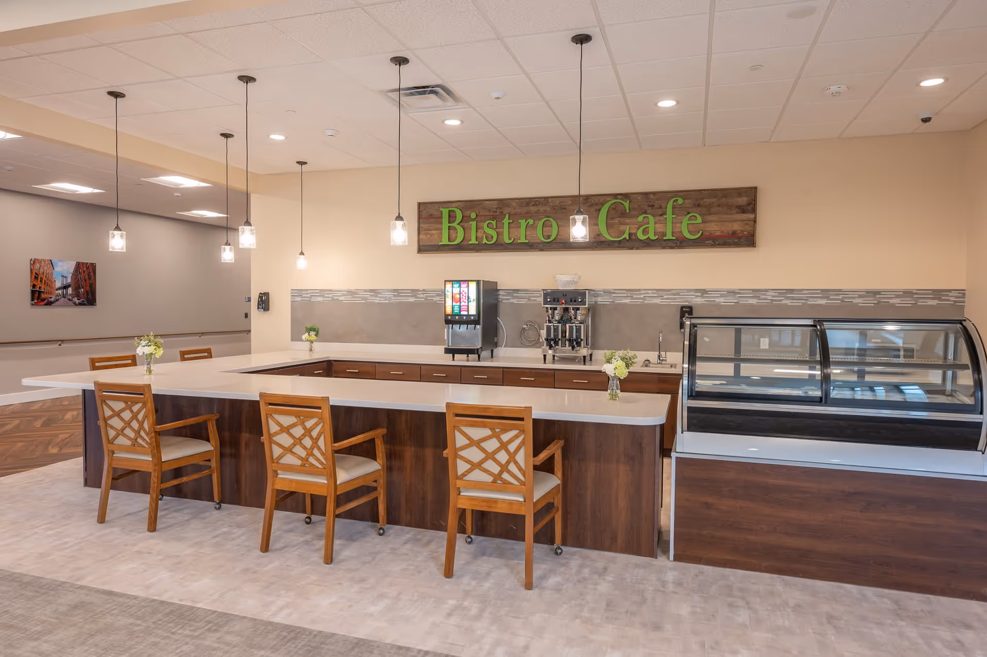 Interior view of a bistro cafe area in a senior living facility with a U-shaped counter, wooden chairs with cushioned seats, hanging pendant lights, a beverage dispenser, a coffee machine, and a glass display case. The wall behind the counter has a wooden sign that reads 'Bistro Cafe' in green letters.