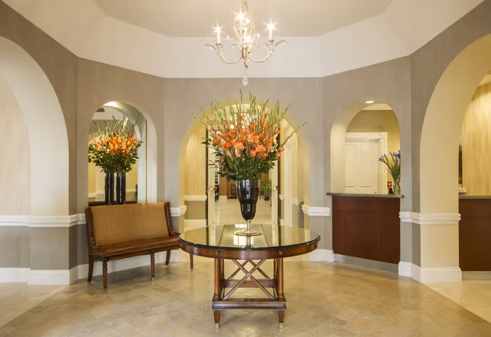 A spacious and elegant lobby area with a round wooden table in the center holding a large vase of orange flowers. The walls have arched openings and are painted in neutral tones with white trim. There is a wooden bench with a cushioned seat to the left and a reception desk to the right. A chandelier hangs from the ceiling, illuminating the space.