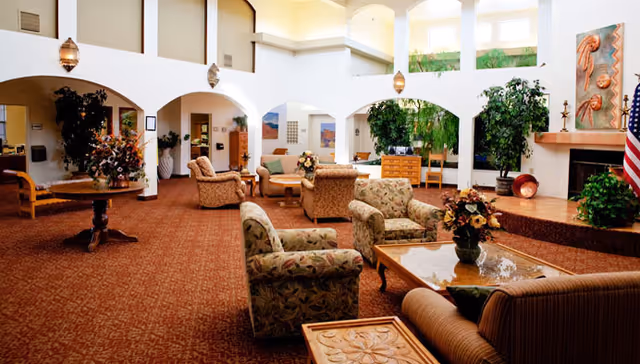 Spacious and well-lit living room area in a senior living facility with multiple upholstered armchairs and sofas arranged around wooden coffee tables. The room features high ceilings with large windows, decorative plants, a fireplace with artwork above it, and an American flag in the corner. The carpet is patterned in a warm tone, and there are archways leading to other parts of the facility.