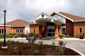 Exterior view of a brick healthcare center building with a covered entrance, a lamp post, and landscaped bushes and plants in front under a cloudy sky.