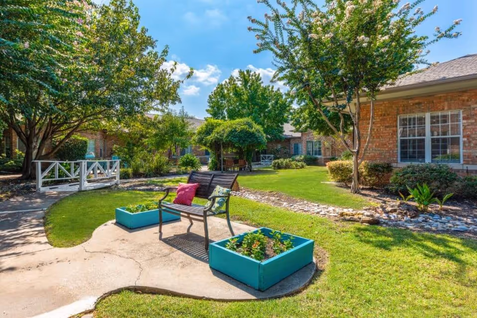 A sunny outdoor garden area at Spring Creek Assisted Living featuring a wooden bench with colorful cushions, two blue planter boxes with plants, a small white footbridge, green grass, trees, and brick buildings in the background under a blue sky with some clouds.