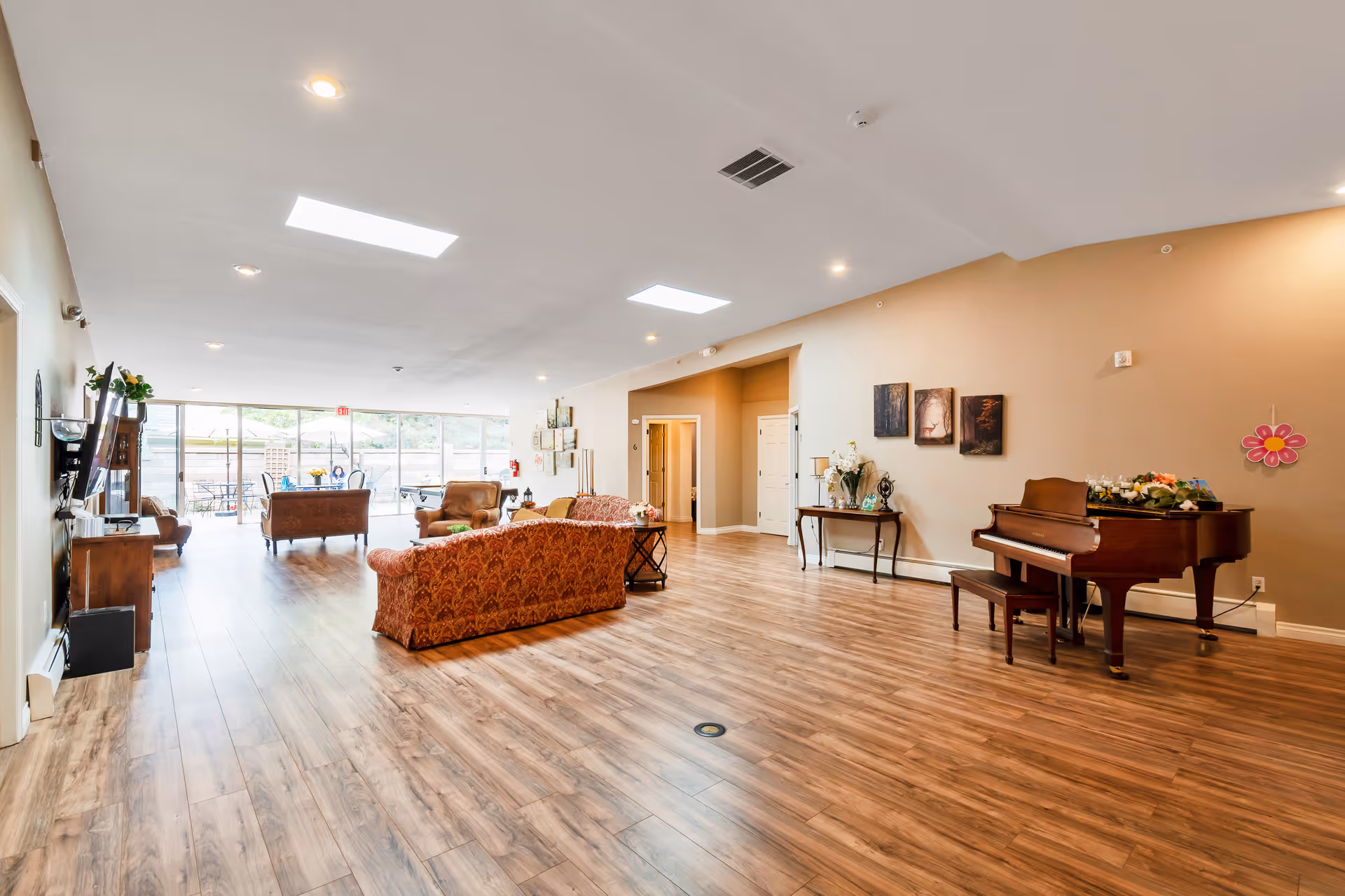 A spacious senior living facility common area with wooden flooring, beige walls, and recessed ceiling lights. The room features a grand piano with a bench on the right side, a small table with decorative items, and several upholstered sofas and armchairs arranged for seating. Large windows and glass doors at the far end allow natural light to fill the space and provide a view of an outdoor patio area with tables and chairs.