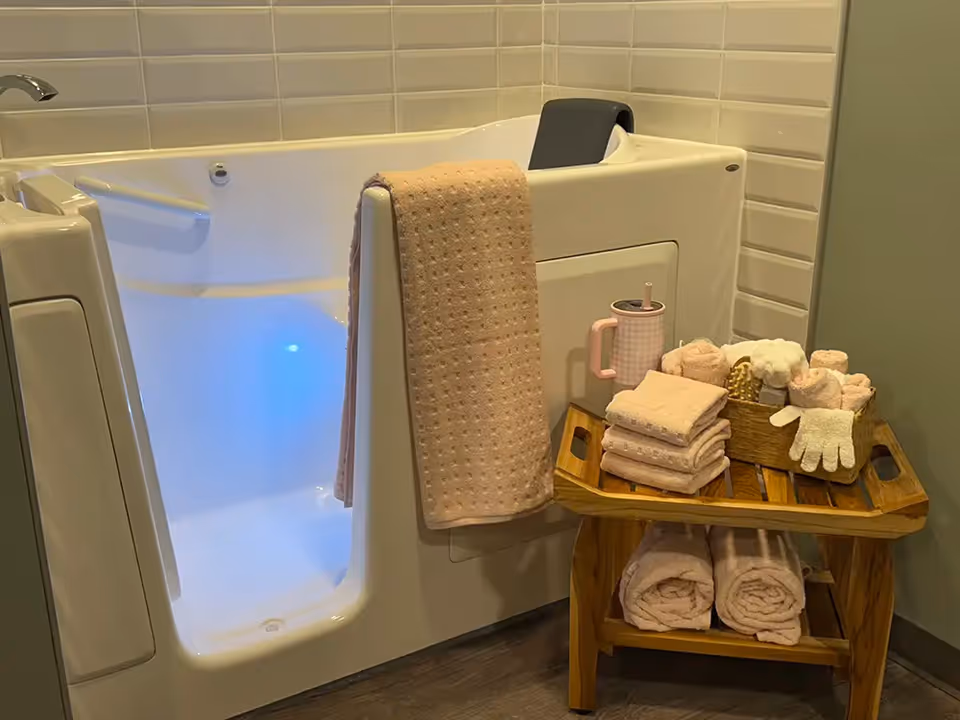 A walk-in bathtub with a door open, illuminated with blue light inside. A beige towel hangs over the side of the tub. Next to the tub is a wooden stool holding neatly folded beige towels, a pink cup with a straw, and a basket containing rolled towels and a white exfoliating glove. The wall behind the tub is tiled with light-colored rectangular tiles.