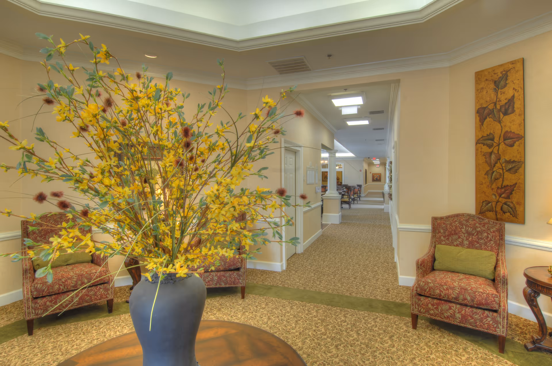A cozy interior hallway in a senior living facility with patterned carpet and beige walls. There are two upholstered armchairs with green cushions on either side of the hallway entrance, a large vase with yellow and brown floral arrangement on a round wooden table in the foreground, and a decorative wall hanging on the right wall. The hallway extends into a dining area with tables and chairs visible in the distance.