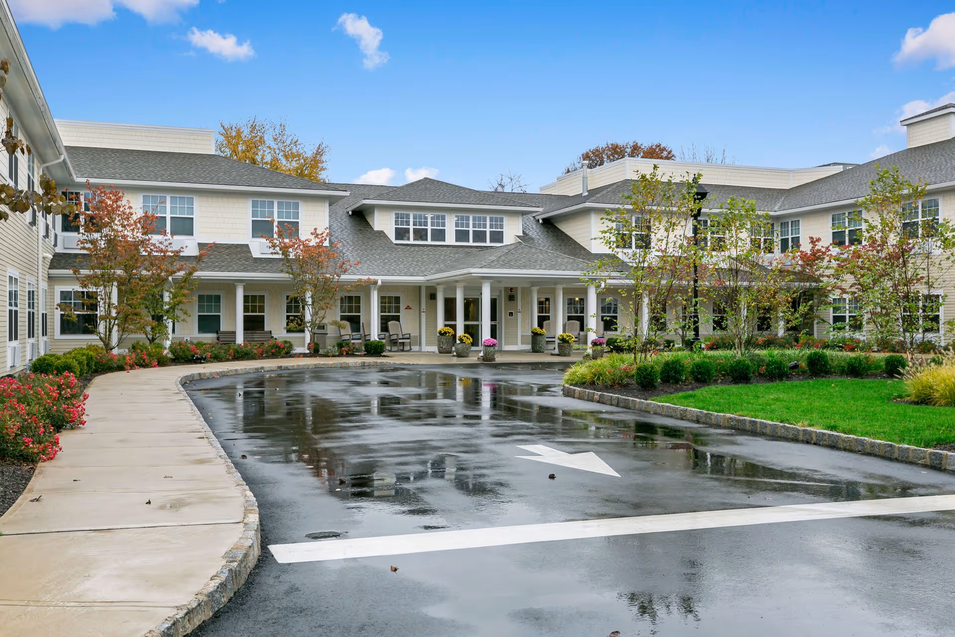 Exterior view of All American Assisted Living at Wareham showing a two-story building with white siding and multiple windows. The entrance is covered by a roof supported by white columns, with a driveway and sidewalk leading up to it. There are landscaped areas with green grass, bushes, and small trees with autumn foliage. The sky is blue with some clouds.