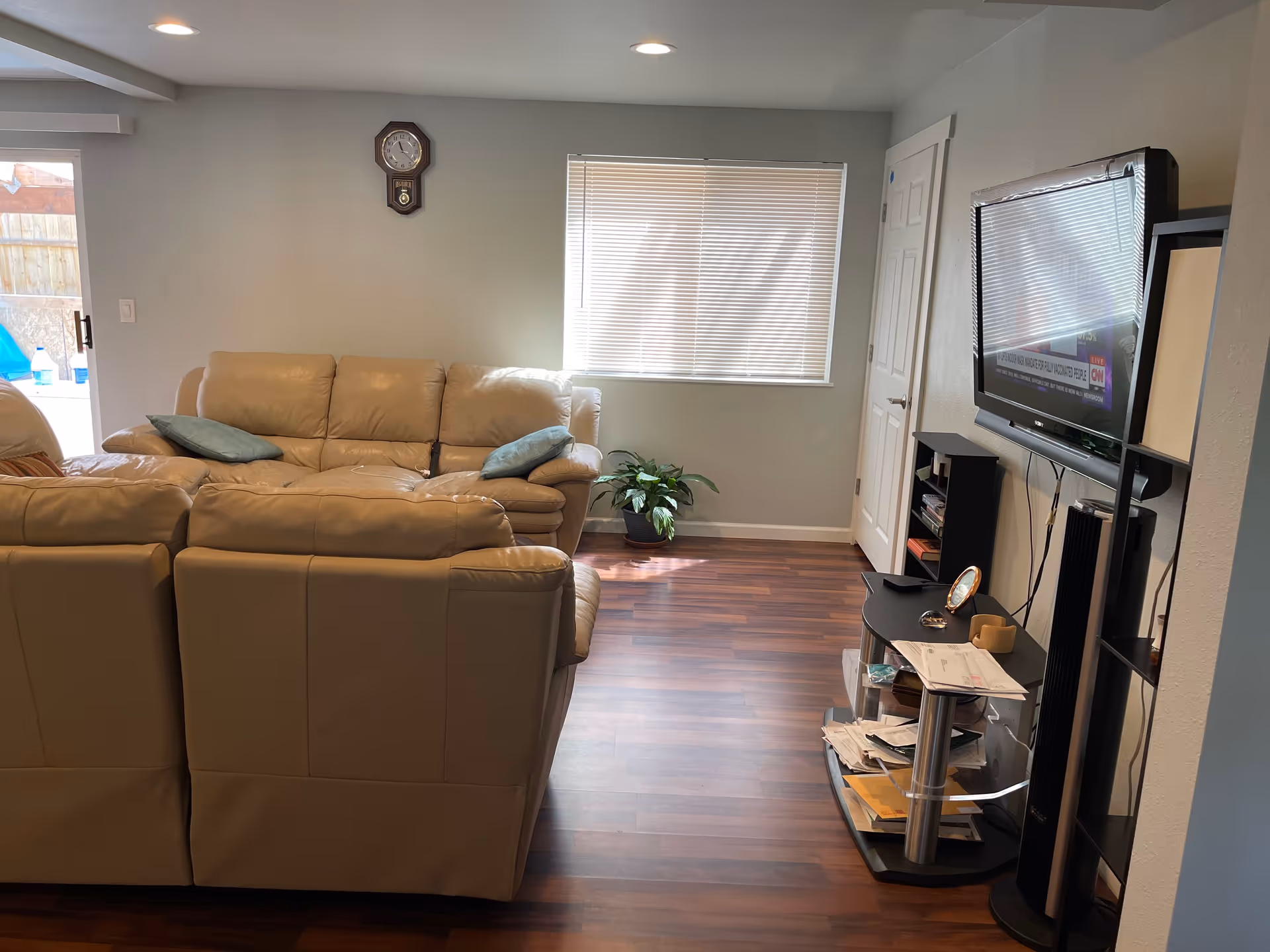 A living room with beige leather sofas, a wall clock, a window with blinds, a potted plant in the corner, and a TV mounted on the wall with a black TV stand below it holding various papers and items. The floor is wooden and there is a door next to the window.