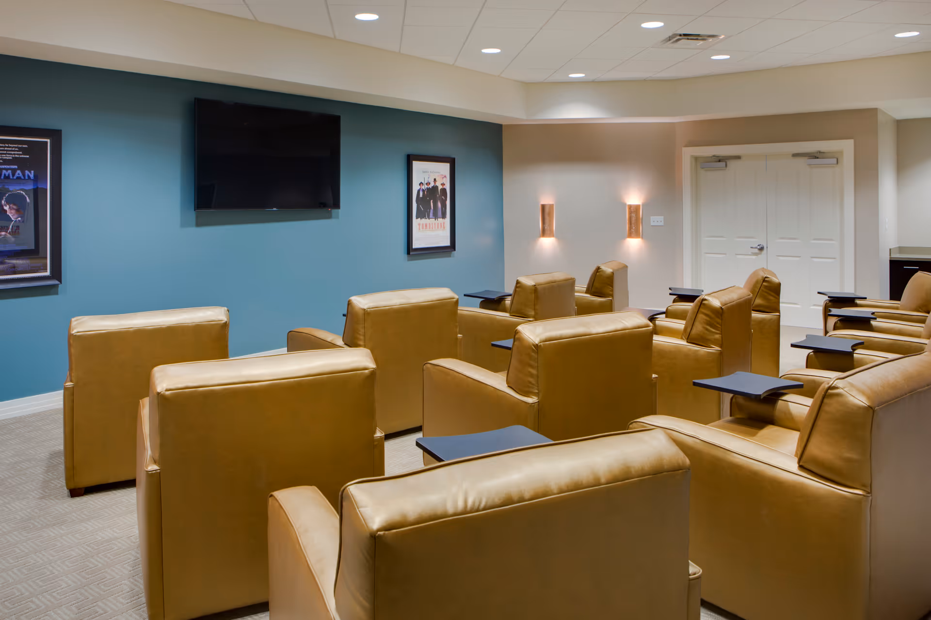 Small theater-style media room with rows of tan leather recliners facing a wall-mounted TV on a teal accent wall.
