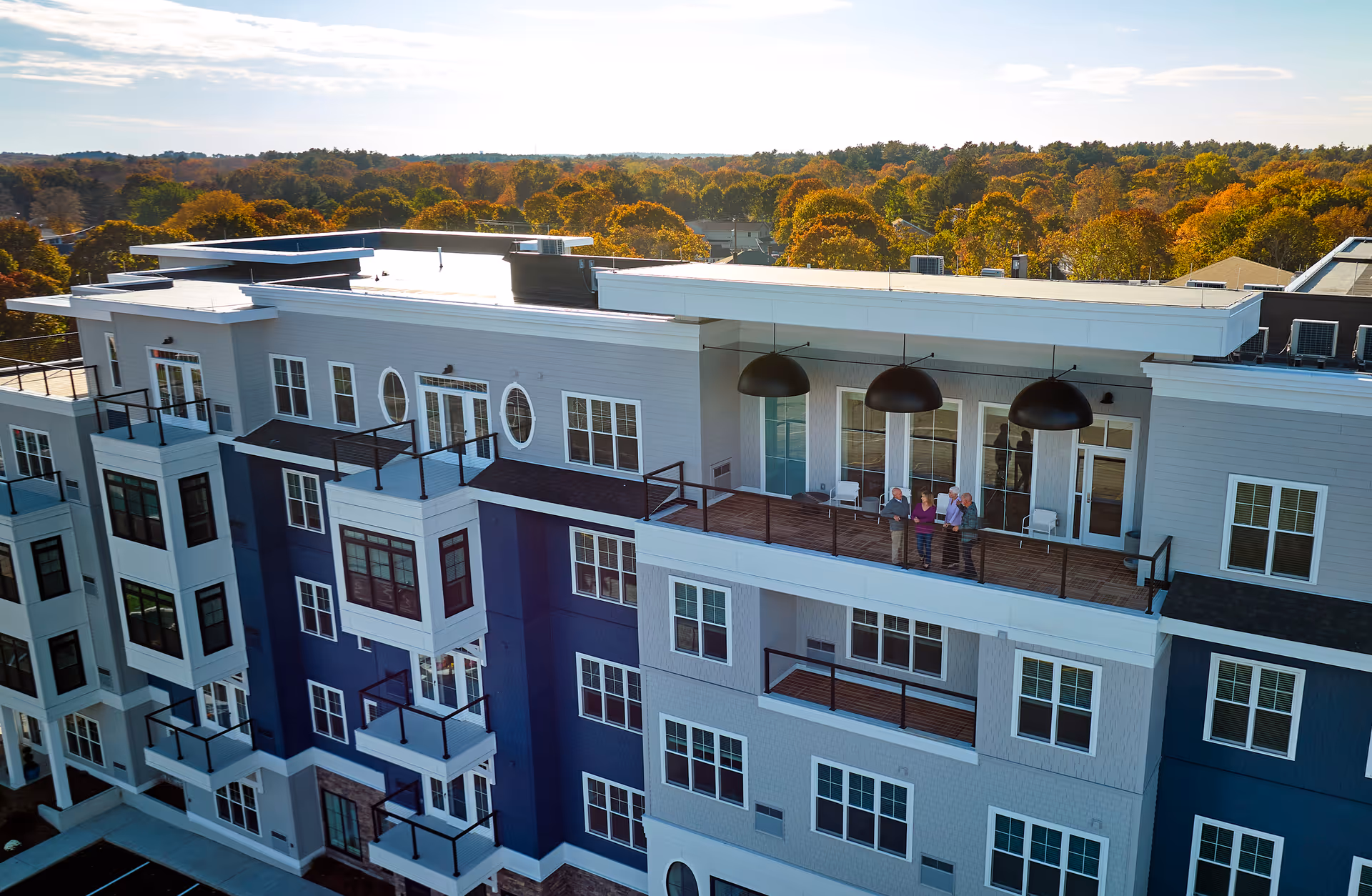 A modern multi-story senior living facility with balconies and a large covered terrace where four elderly people are standing and conversing. The building exterior features a combination of light gray and blue siding with multiple windows. The background shows a scenic view of trees with autumn foliage under a partly cloudy sky.