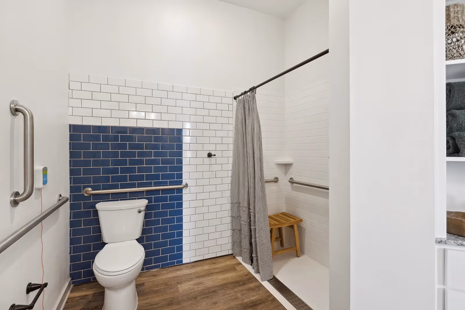 A modern bathroom featuring a white toilet against a wall with white and blue subway tiles. There are stainless steel grab bars on the walls for accessibility. To the right, there is a walk-in shower with a gray striped curtain, white tiled walls, a wooden shower stool, and additional grab bars. The floor is wood-style laminate.