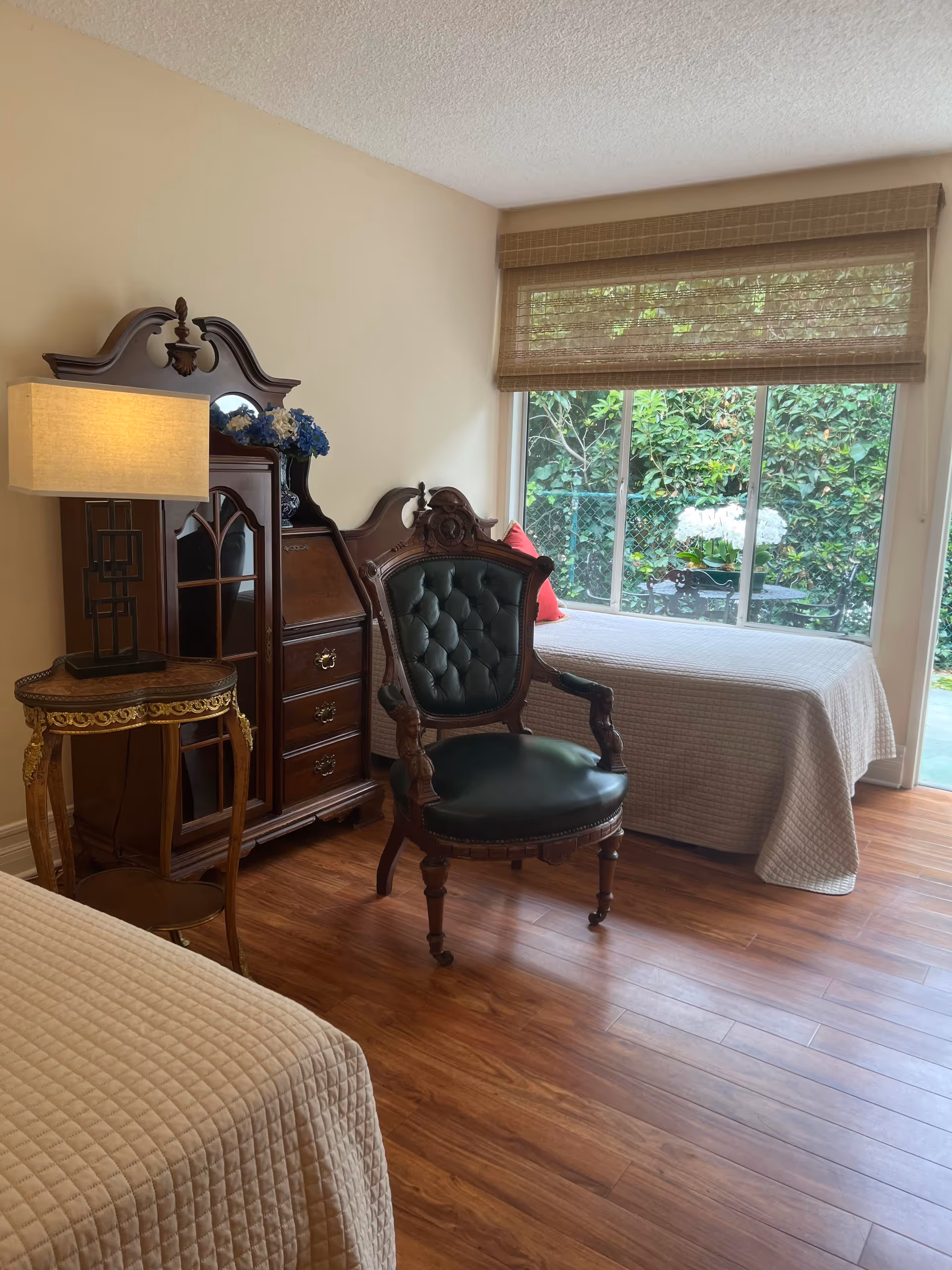 A bedroom with two twin beds covered in beige quilted bedspreads, a vintage wooden armchair with dark green leather upholstery, a wooden cabinet with drawers and a glass door, a small round wooden side table with a lamp, and a large window with bamboo blinds showing greenery outside.
